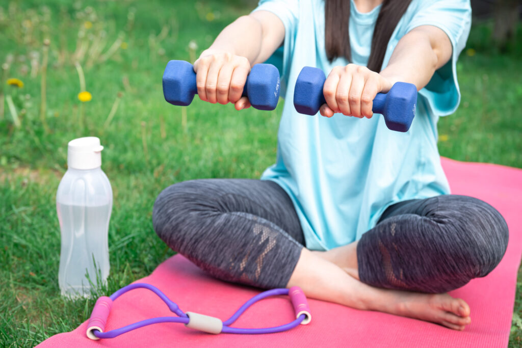 Sportive young woman stretching with dumbbells, doing fitness exercises in green park.