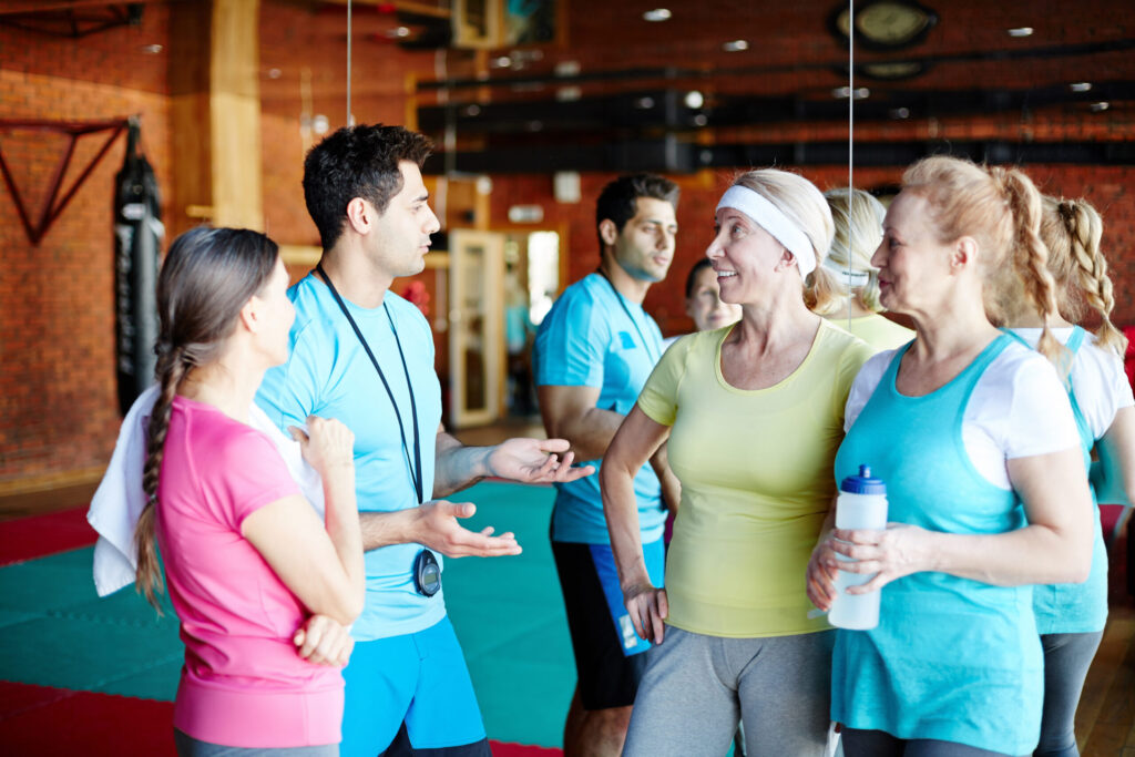 Young trainer explaining some training moments to team of three aged women before workout in gym